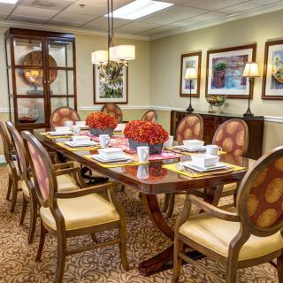 A well-decorated dining or conference room with a large wooden table set for a meal and adorned with flowers, surrounded by chairs and illuminated by a chandelier.