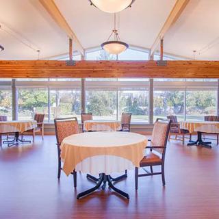 A spacious dining room with large windows and wooden flooring, featuring multiple tables and chairs.