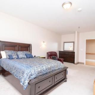 A spacious bedroom with a large bed, dresser, and chair, featuring neutral-colored walls and carpeting.