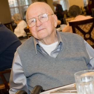 An elderly man in glasses sits in a dining area, wearing a grey vest over a shirt.
