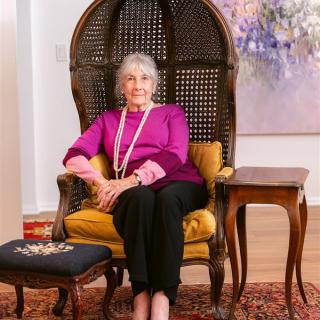 An elderly woman sits comfortably on a chair in a well-decorated living room.