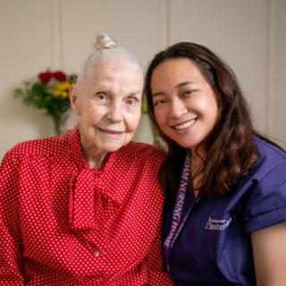 An elderly woman in a red dress smiling with a caregiver beside her inside a room.