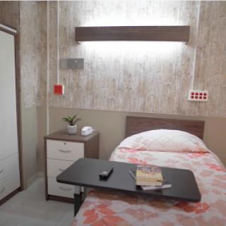A clean and minimalist hospital room featuring a single bed, nightstand, wardrobe, and a small table.