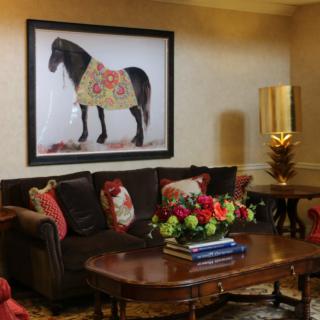 A cozy living room featuring a dark sofa adorned with pillows, a wooden coffee table with a colorful flower arrangement, and a large artwork of a horse on the wall.