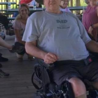 A man in a wheelchair is at an outdoor gathering with people in the background.