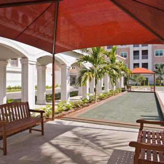 A serene outdoor patio featuring benches, a bocce court, and a red canopy with palm trees lining the walkway.