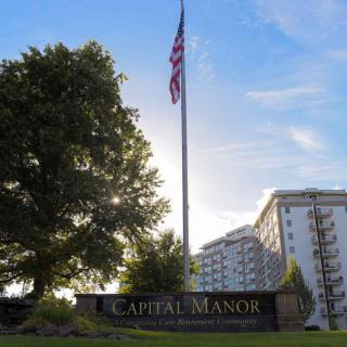 A large flagpole with an American flag stands in front of Capital Manor, a multi-story building surrounded by trees under a clear sky.