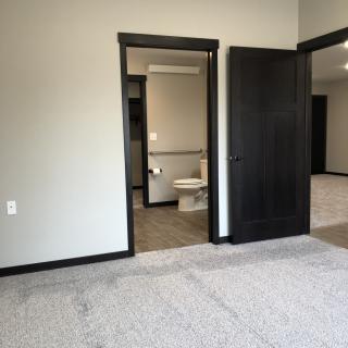 A modern bedroom with an adjoining bathroom, featuring carpeted flooring and dark-colored door frames.