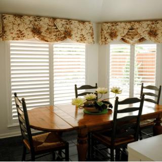 A cozy dining room with a wooden table and chairs, featuring floral curtains over large windows.