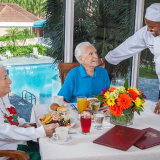 A chef interacting with two seniors enjoying a meal by the pool.