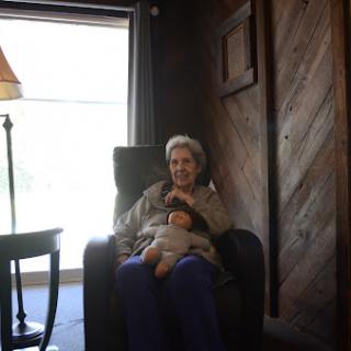 An elderly woman sitting comfortably in a chair, holding a doll, with natural light streaming through a nearby window.