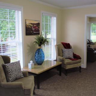 A cozy living room interior with soft chairs, a table with a decorative vase, and large windows.