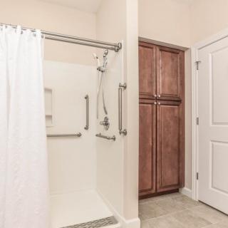 A bathroom featuring a shower with a white curtain and safety grab bars, alongside a wooden storage cabinet and a nearby door.