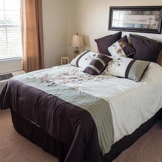 A well-decorated bedroom featuring a neatly made bed with patterned bedding and pillows, complemented by a bedside lamp and a window providing natural light.