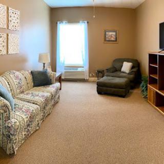 A cozy living room featuring a patterned sofa, armchair, television, and a window letting in natural light.