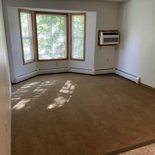 An empty room with a carpeted floor, featuring a bay window and an air conditioner.