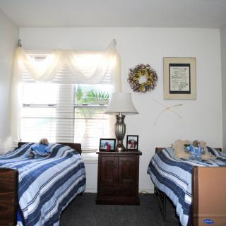 A cozy bedroom with twin beds featuring blue and white striped bedding, separated by a wooden nightstand with a table lamp. The room is decorated with framed pictures and receives natural light through the window.
