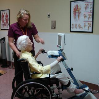 An elderly person in a wheelchair using exercise equipment with assistance from a caregiver.