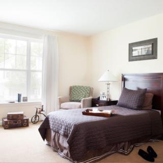 A cozy bedroom with a large window, featuring a neatly made bed, a chair, and decorative elements.