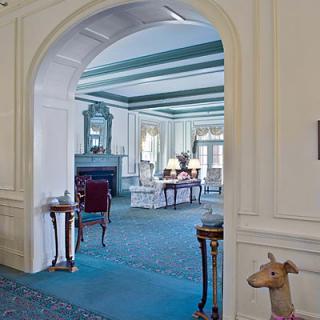 A classic, elegantly decorated room with a view through an archway, showcasing period furniture and a large fireplace.