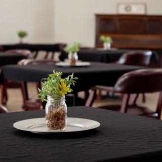 A dining area with tables covered in black cloth, each adorned with a jar centerpiece containing greenery.