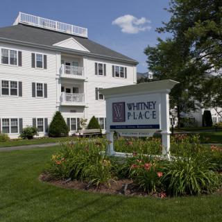 A white residential building with a sign labeled 'Whitney Place' surrounded by well-maintained landscaping and flowers.