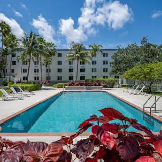 A serene swimming pool surrounded by lush greenery and an apartment complex under a clear blue sky.