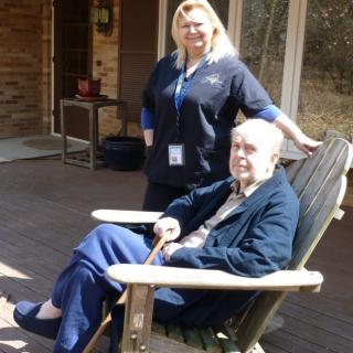 An elderly person sitting in a patio chair with a caregiver standing nearby.