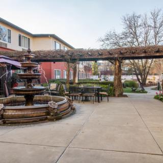 A tranquil courtyard featuring a central fountain, surrounded by outdoor seating and a pergola, with garden views and an adjacent building.