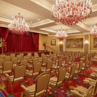 An elegant conference room with rows of chairs facing a stage, adorned with chandeliers and red curtains.