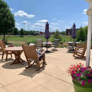 A sunny patio area with wooden chairs and tables, surrounded by greenery and colorful flowers.