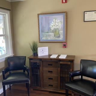 A cozy waiting room featuring two chairs, a side cabinet with paperwork, and a decorative painting on the wall.