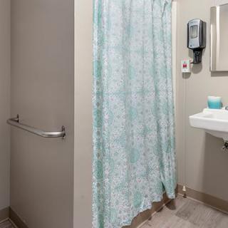 A bathroom featuring a shower with a patterned curtain, a sink with a mirror above it, and a soap dispenser on the wall.