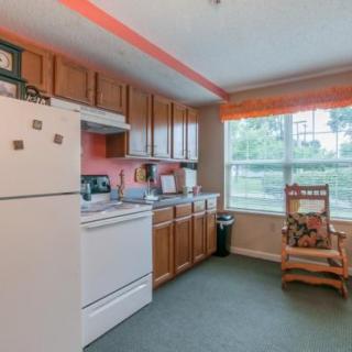 A cozy kitchen with wooden cabinets, a white fridge and stove, a large window, and a rocking chair.