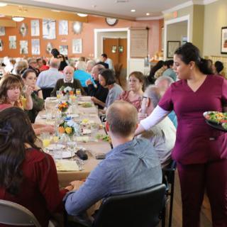 A communal dining scene with people enjoying a meal together, as a server attends to the guests.