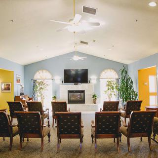 A spacious living room with arranged chairs facing a television above a fireplace, complemented by large windows and indoor plants.