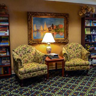 A cozy living room area with two armchairs, a lamp on a side table, bookshelves, and a painting on the wall.
