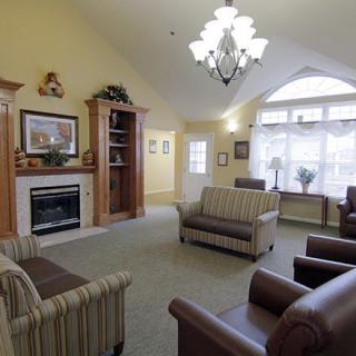 A cozy living room with a fireplace, sofas, and armchairs under a chandelier, featuring large windows for natural light.