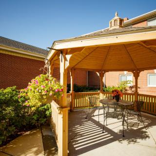 A sunny day view of a wooden gazebo with patio furniture and hanging plants, set against a brick building backdrop.