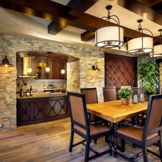 A modern dining room featuring a wooden table and chandeliers, with an open kitchen and stone wall.