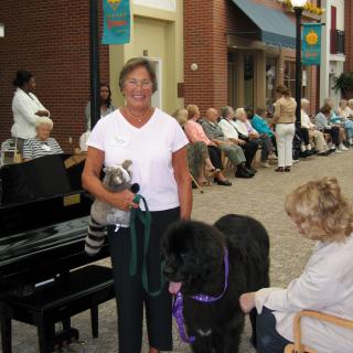 A person stands with a dog on a street while another person sits nearby, with a crowd in the background.