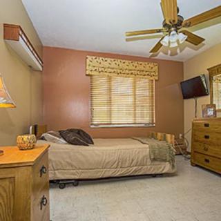 A cozy bedroom with neutral tones, featuring a single bed, wooden dresser, ceiling fan, and a window with curtains.