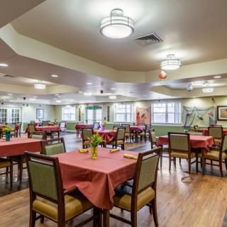 A brightly lit dining room with neatly arranged tables and chairs, featuring modern light fixtures and large windows.