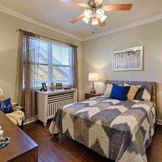 A cozy and well-decorated bedroom with a patterned quilt, ceiling fan, and natural lighting.