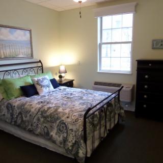 A cozy bedroom featuring a neatly made bed with patterned bedding, a lamp on the bedside table, and a window bringing in natural light.