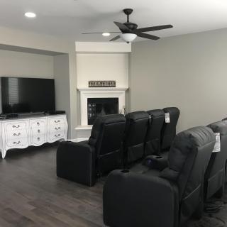 A cozy home theater setup with multiple recliners facing a large television, complemented by a fireplace and ceiling fan.