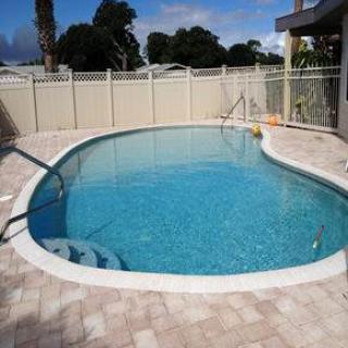 A backyard swimming pool surrounded by a paved area with a fence and trees in the background.