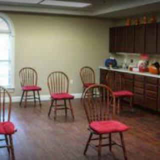 A room with wooden chairs arranged in a circle and a kitchenette in the background.