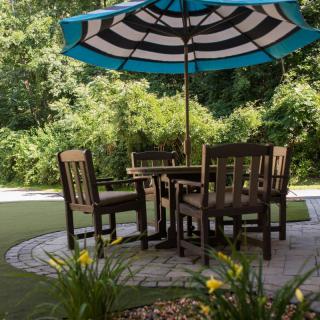 A cozy outdoor patio setup with a table and chairs under a large striped umbrella, surrounded by greenery.