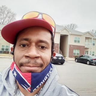 A man wearing a cap and mask takes a cheerful selfie outside an apartment building.